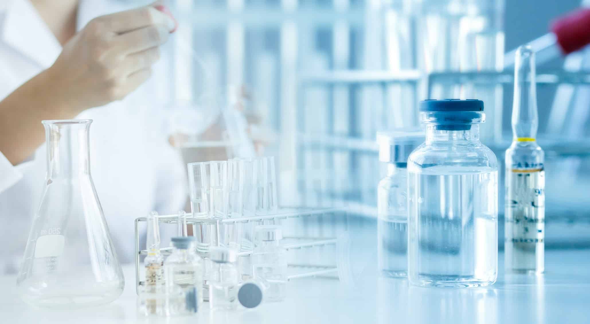 Sealed drug vials, glass ampoules, a test tube rack and an Erlenmeyer flask on a lab bench, with a gloved researcher working out of focus behind them.