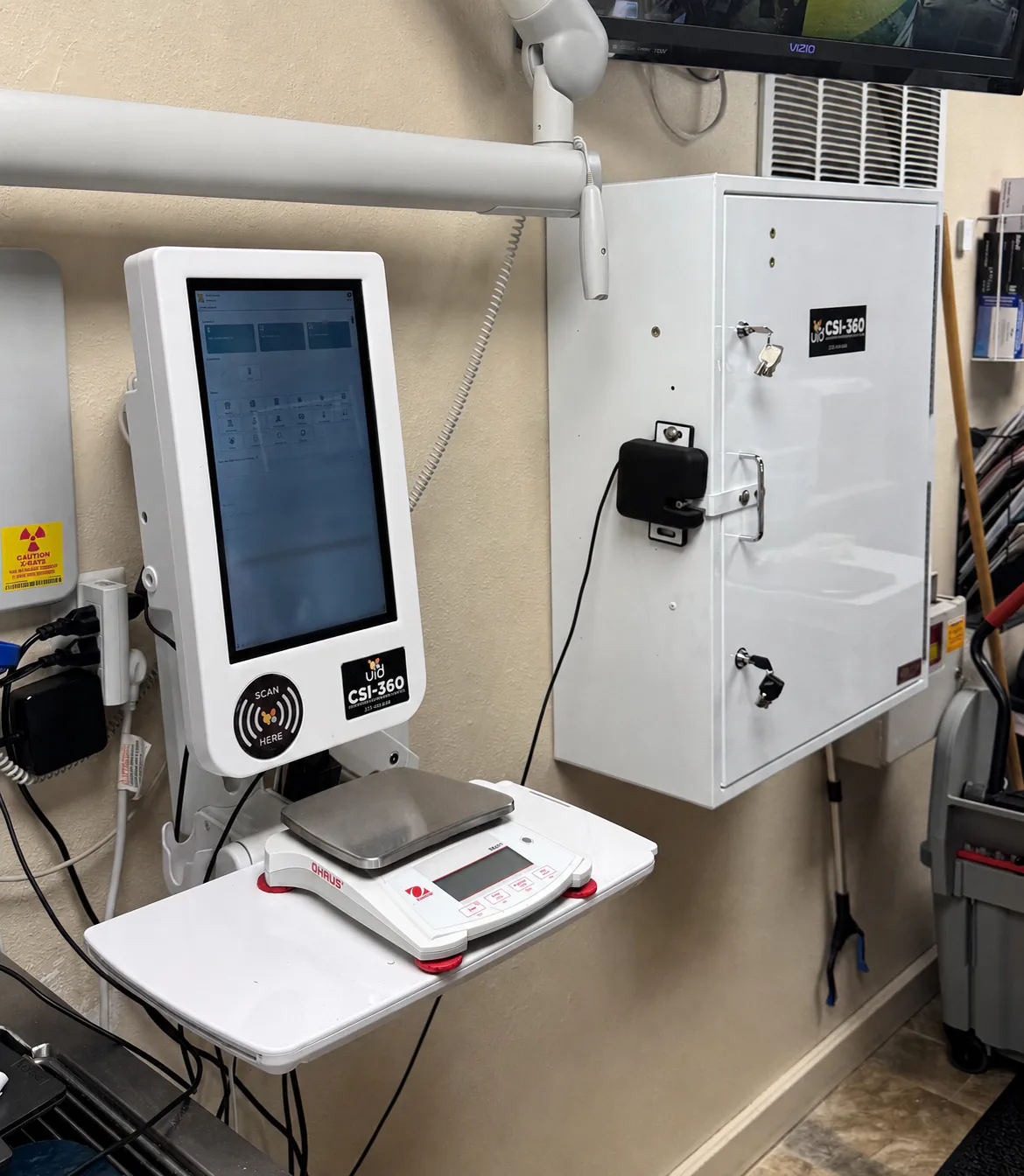 CSI-360 touchscreen and digital scale beside a wall-mounted white controlled-substance cabinet in a veterinary treatment area