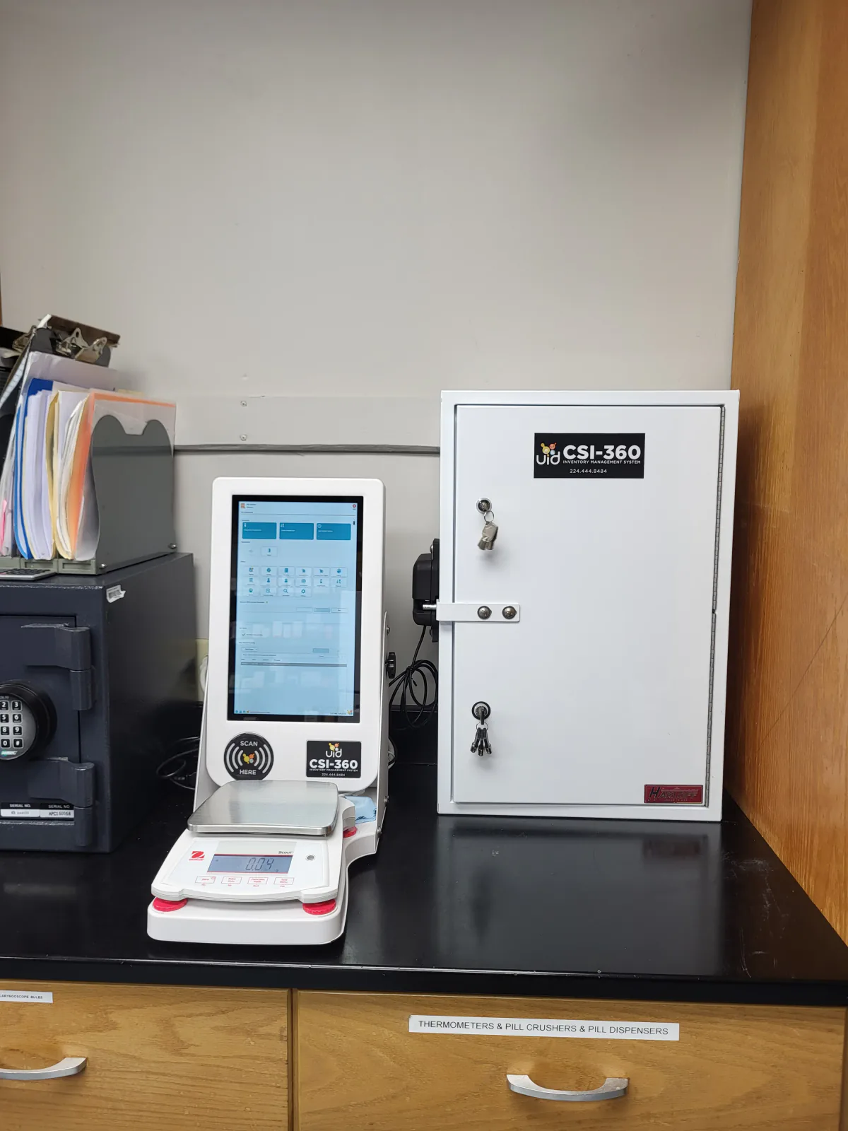 CSI-360 touchscreen and digital scale on a clinic counter beside a white cabinet and an existing black keypad safe